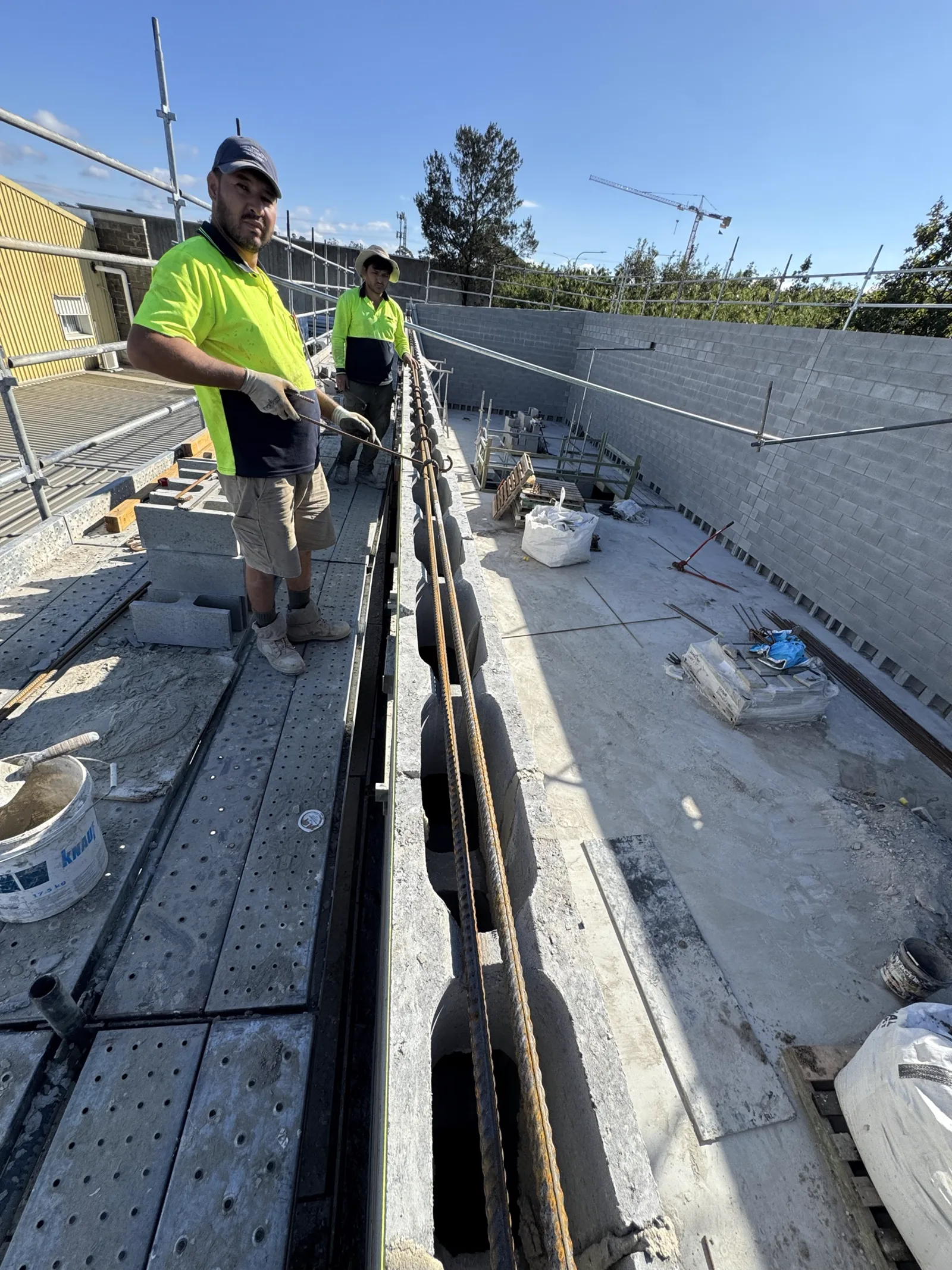 Bricklayers threading reinforcement bar into block cores ahead of core filling on a commercial site.