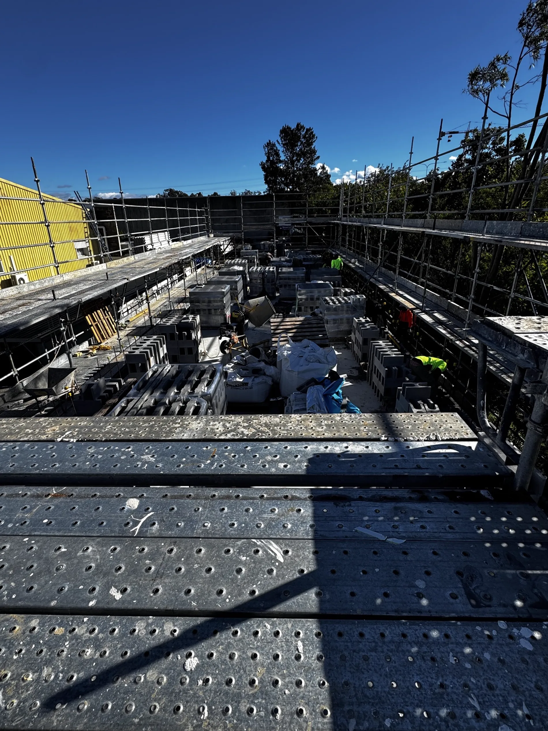 Large commercial construction site with multi-level perimeter scaffold and bulk pallets of concrete block staged across the floor plate.